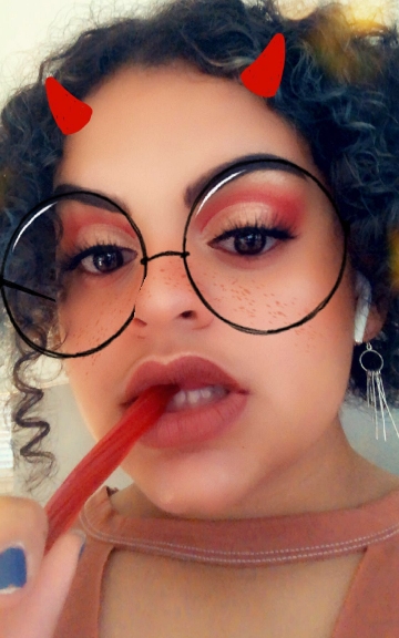 Mya in 2019 with a red Twizzler, red lipstick, red eye makeup, & Snapchat's little red horns & big honking glasses.